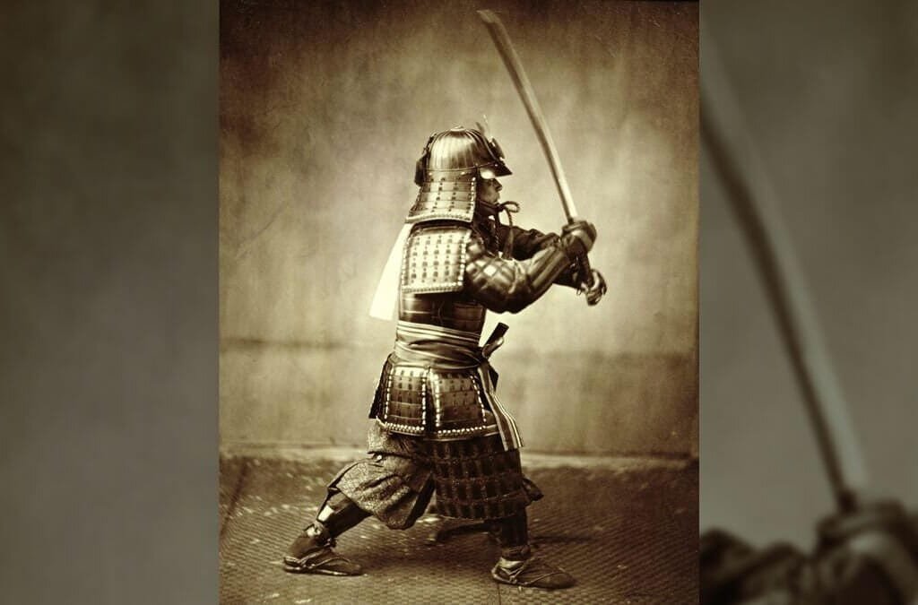 An old black-and-white image of a man standing in profile. He is dressed in samurai armor and is holding a sword upright, as if ready to strike.